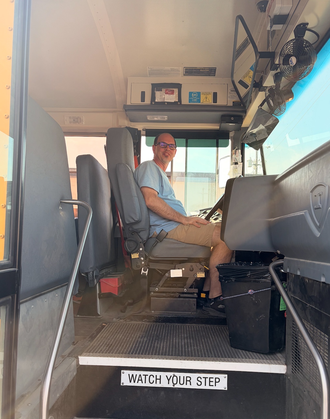 A man in glasses sits in the driver's seat of a school bus, smiling.