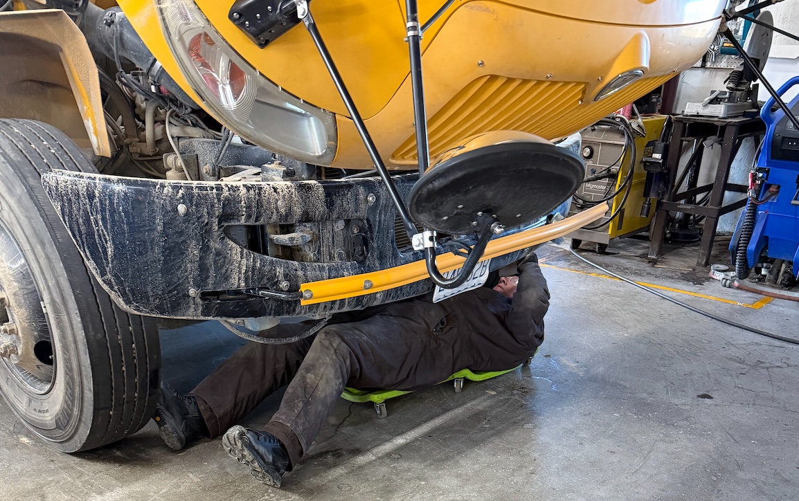 A mechanic lies on a creeper, while inspecting underneath the front of a yellow school bus.