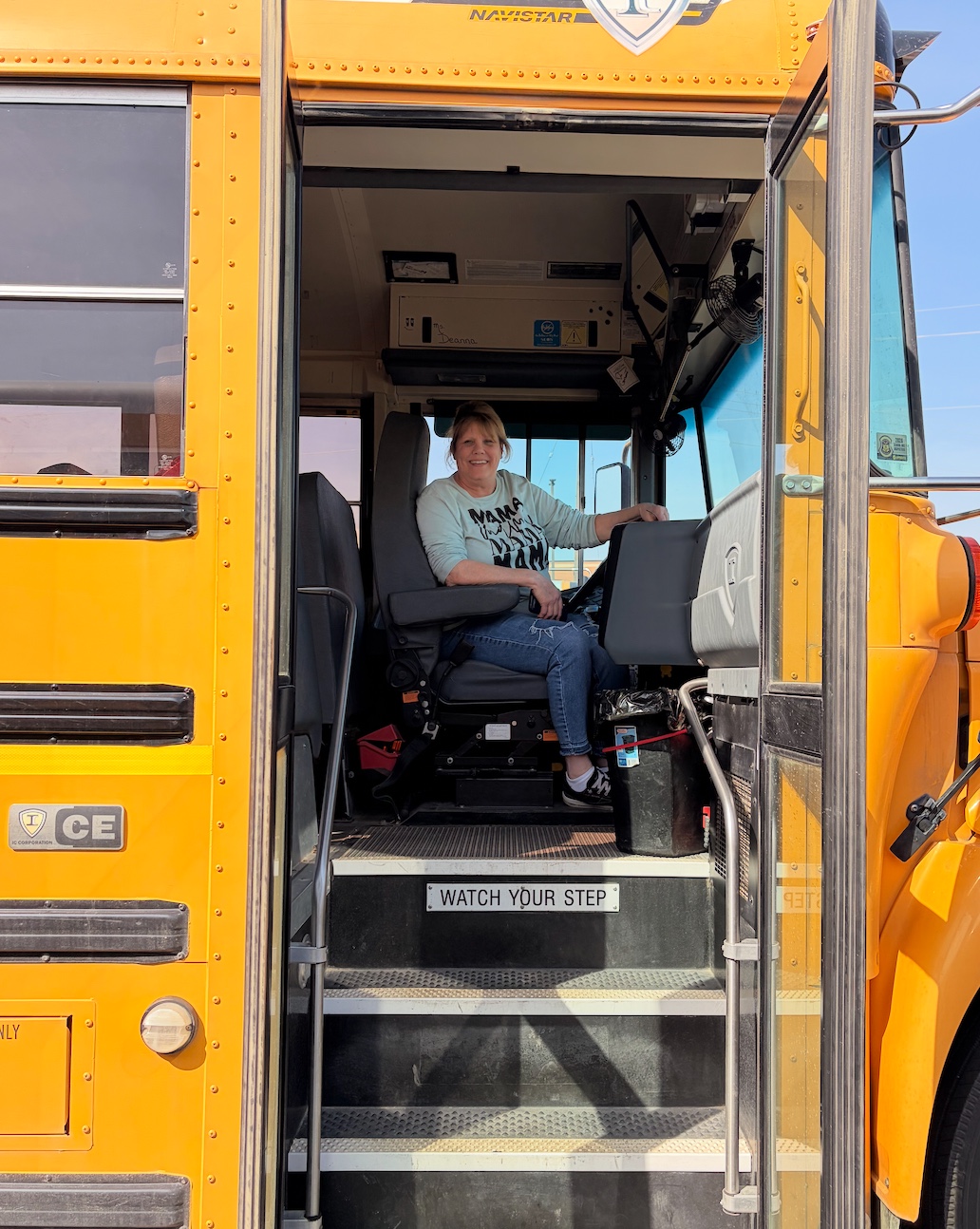 A woman smiles while sitting in the driver's seat of a yellow school bus.