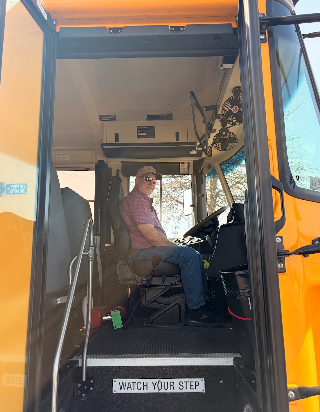 A man in a cap and sunglasses sits in the driver's seat of a yellow school bus.