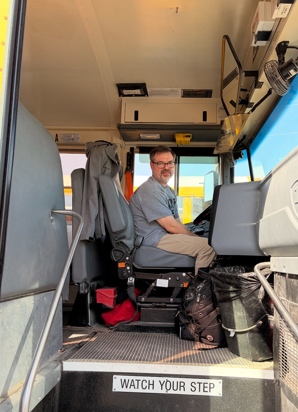 A man in glasses sits in the driver's seat of a school bus, looking towards the camera.