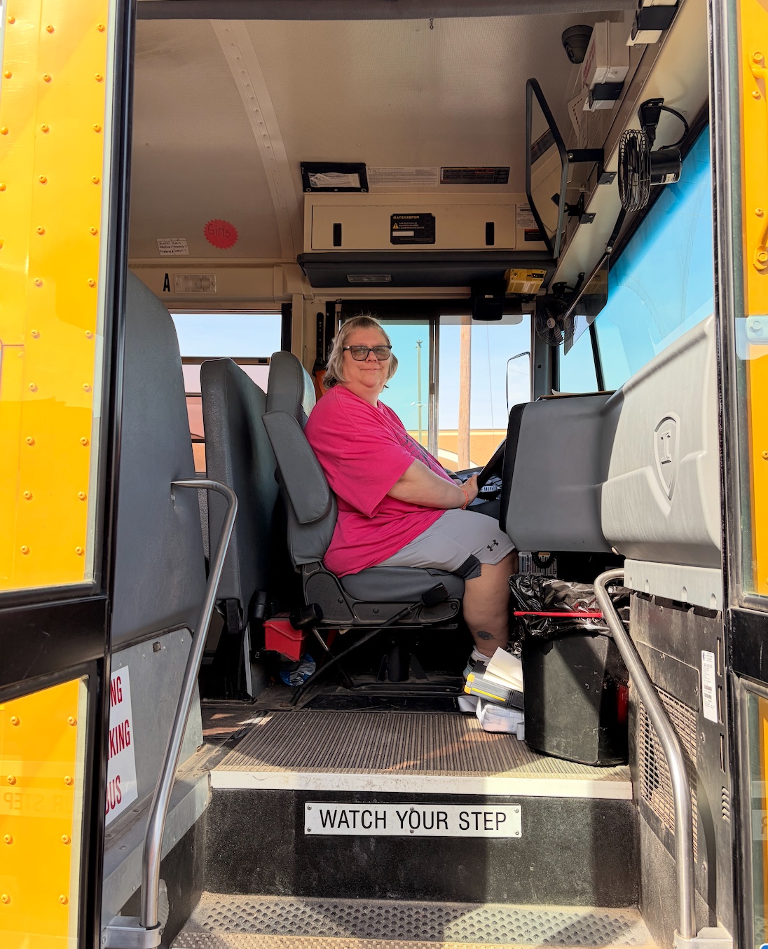 A woman in a pink shirt sits in the driver's seat of a yellow school bus.