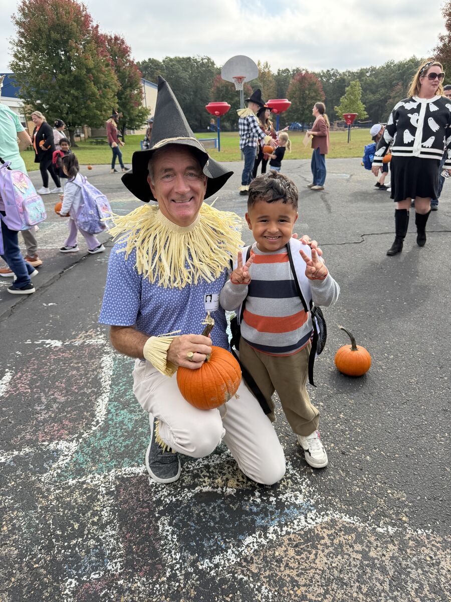 A smiling adult dressed as a scarecrow holds a pumpkin while posing with a young child flashing a peace sign outdoors.