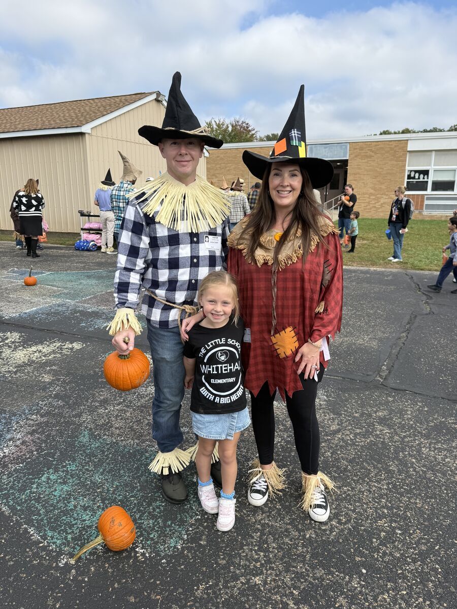 A family poses outside in scarecrow costumes, holding pumpkins on a paved area near a school building.