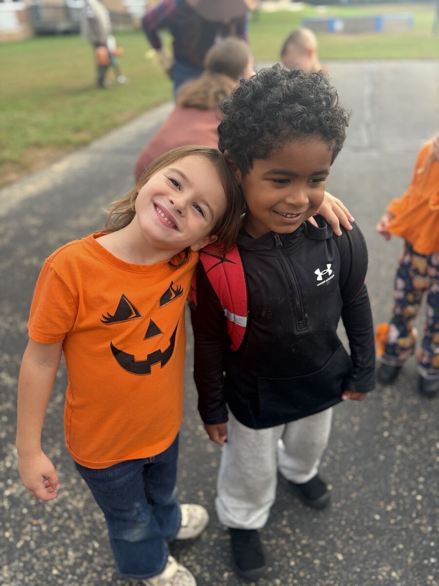 Two smiling children stand close together outdoors, with one leaning their head affectionately on the other's shoulder.