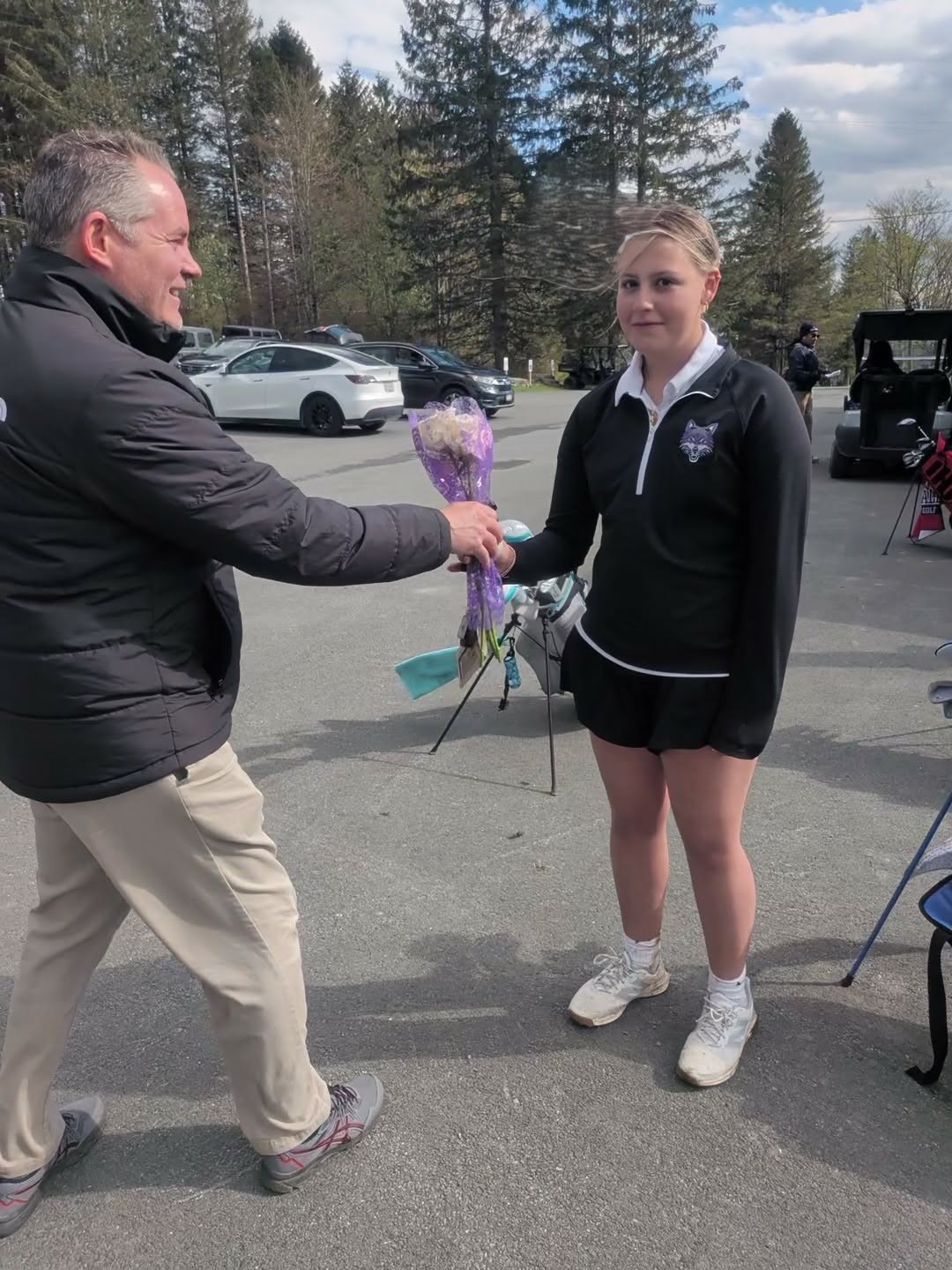 A man in a black jacket hands a bouquet of flowers to a young woman in golf attire.