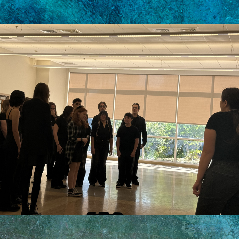 A group of young people in black attire stand in a bright room, facing a person with their back to the camera.