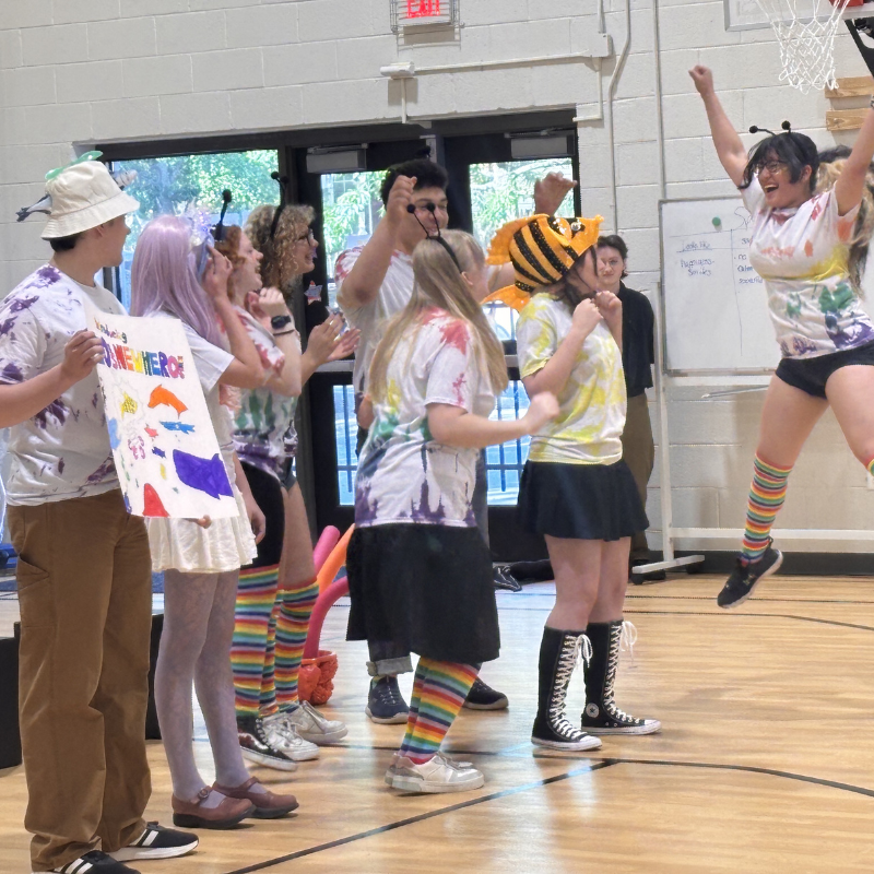 A group of students in colorful tie-dye shirts and fun hats celebrate in a gymnasium.