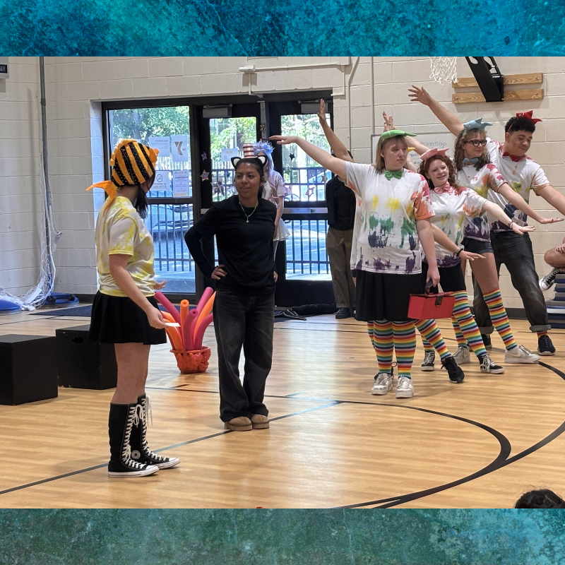 A group of young people in colorful costumes and headwear pose on a wooden floor, possibly during a performance or rehearsal.