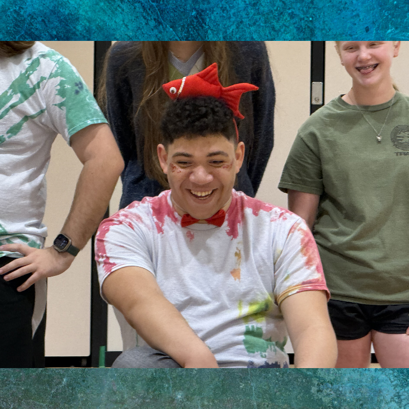 A young person with a red fish headband and bow tie smiles brightly, adorned with glitter.