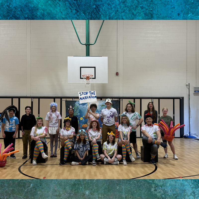 A group of young people in colorful costumes pose in a gymnasium, holding a sign that says "STOP THE NARRATION".
