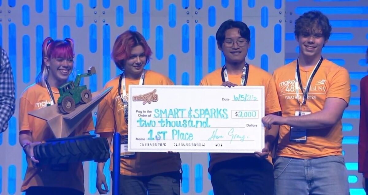 Four young people in orange shirts hold a large check for $2,000, awarded to "SMART & SPARKS" for 1st Place.