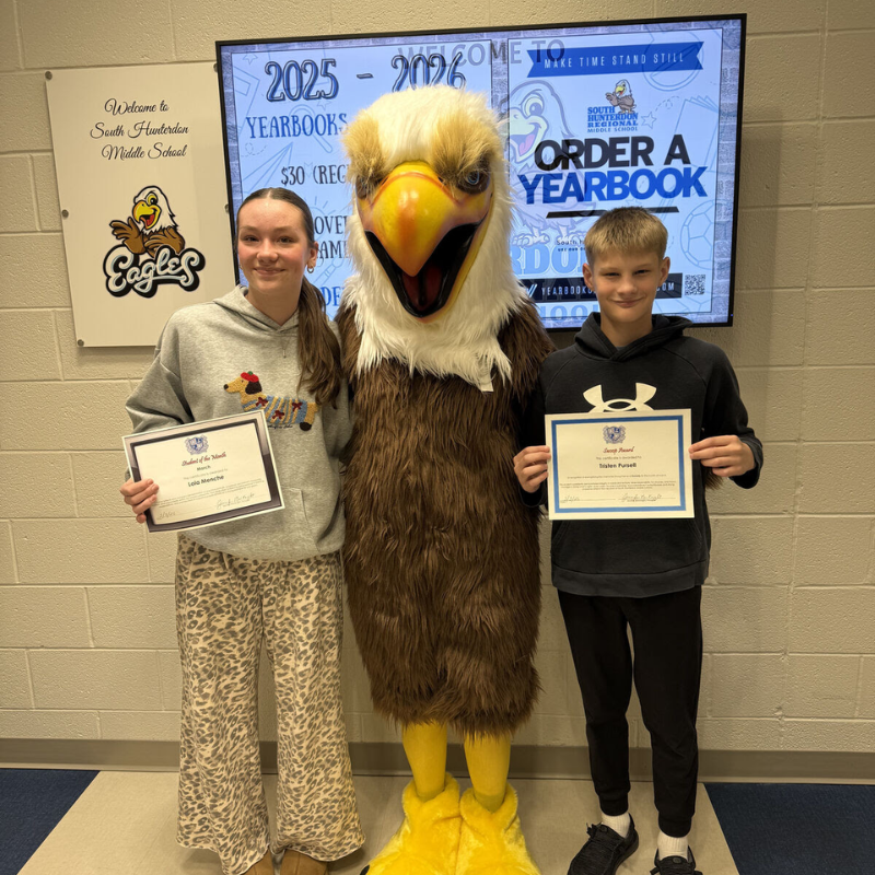 Two students and an eagle mascot pose with awards in front of a school announcement board.