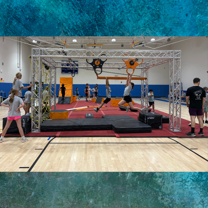 Children practice on an indoor ninja warrior obstacle course with hanging bars and climbing structures.