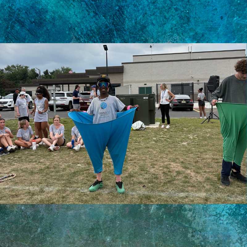 A young person in a grey t-shirt with 'QUALLCO' logo holds up oversized blue pants.