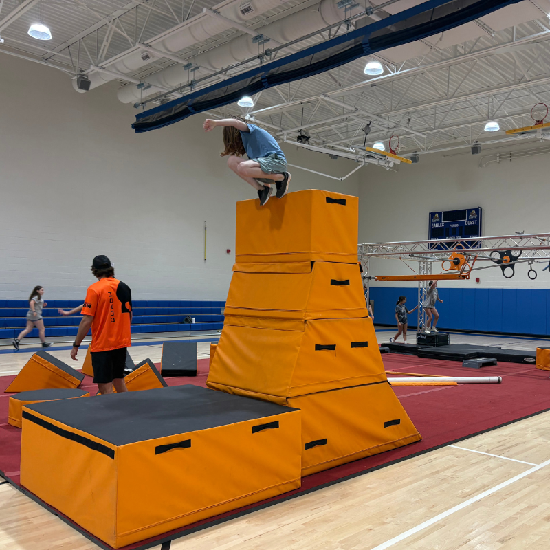 A young person leaps from a tall stack of orange padded blocks in a gymnasium obstacle course.