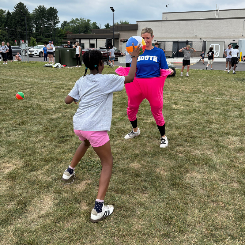 A young girl in a grey shirt and pink shorts throws a ball towards a woman in a blue shirt and oversized pink pants.
