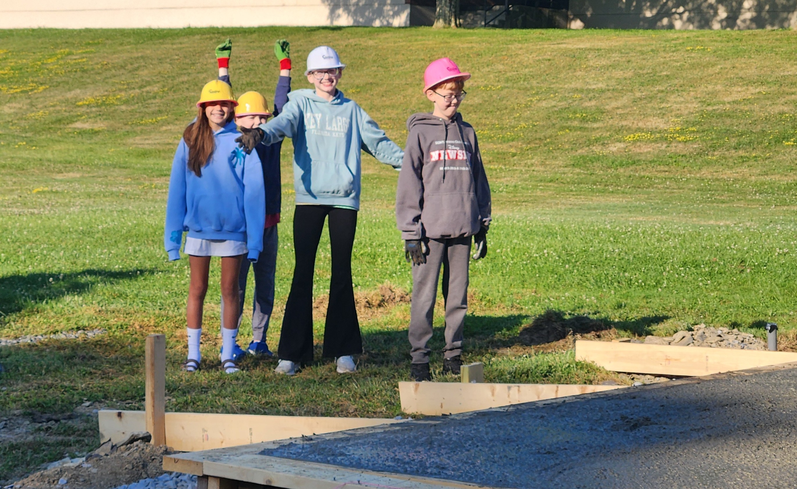 Four children wearing hard hats pose together near a freshly poured concrete slab outdoors.