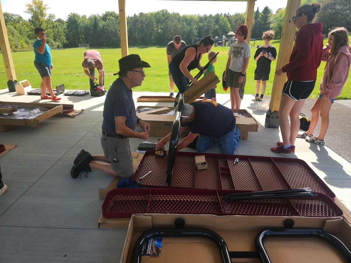 A diverse group of people assemble picnic tables outdoors under a pavilion on a sunny day.