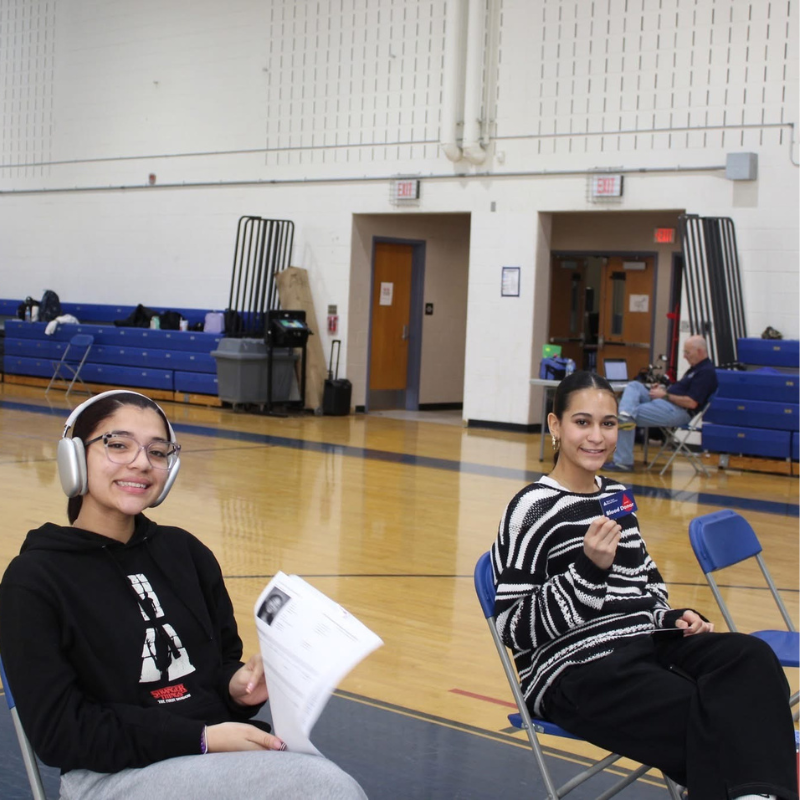 Two young women sit in chairs in a gymnasium, one wearing headphones and holding papers, the other holding a badge.