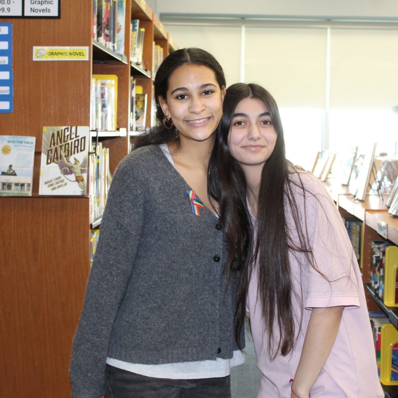 Two young women smile at the camera in a library aisle, with bookshelves in the background.