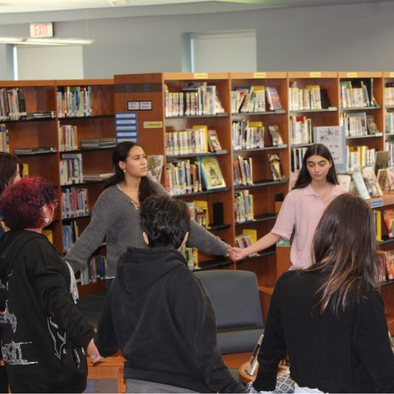 A group of young people hold hands in a circle in a library, surrounded by bookshelves.