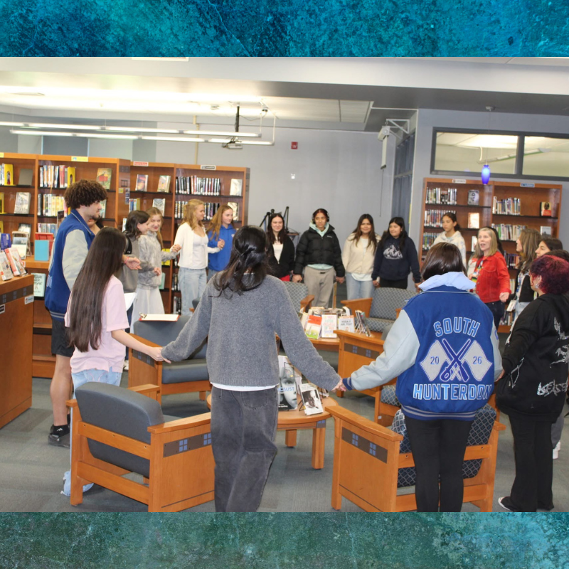 A group of students stand in a circle, holding hands in a library setting.