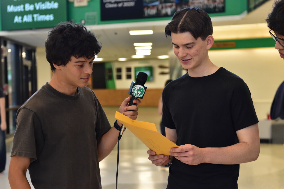 A student holds a microphone with a "DALERCAST" logo while interviewing another student holding a yellow folder.