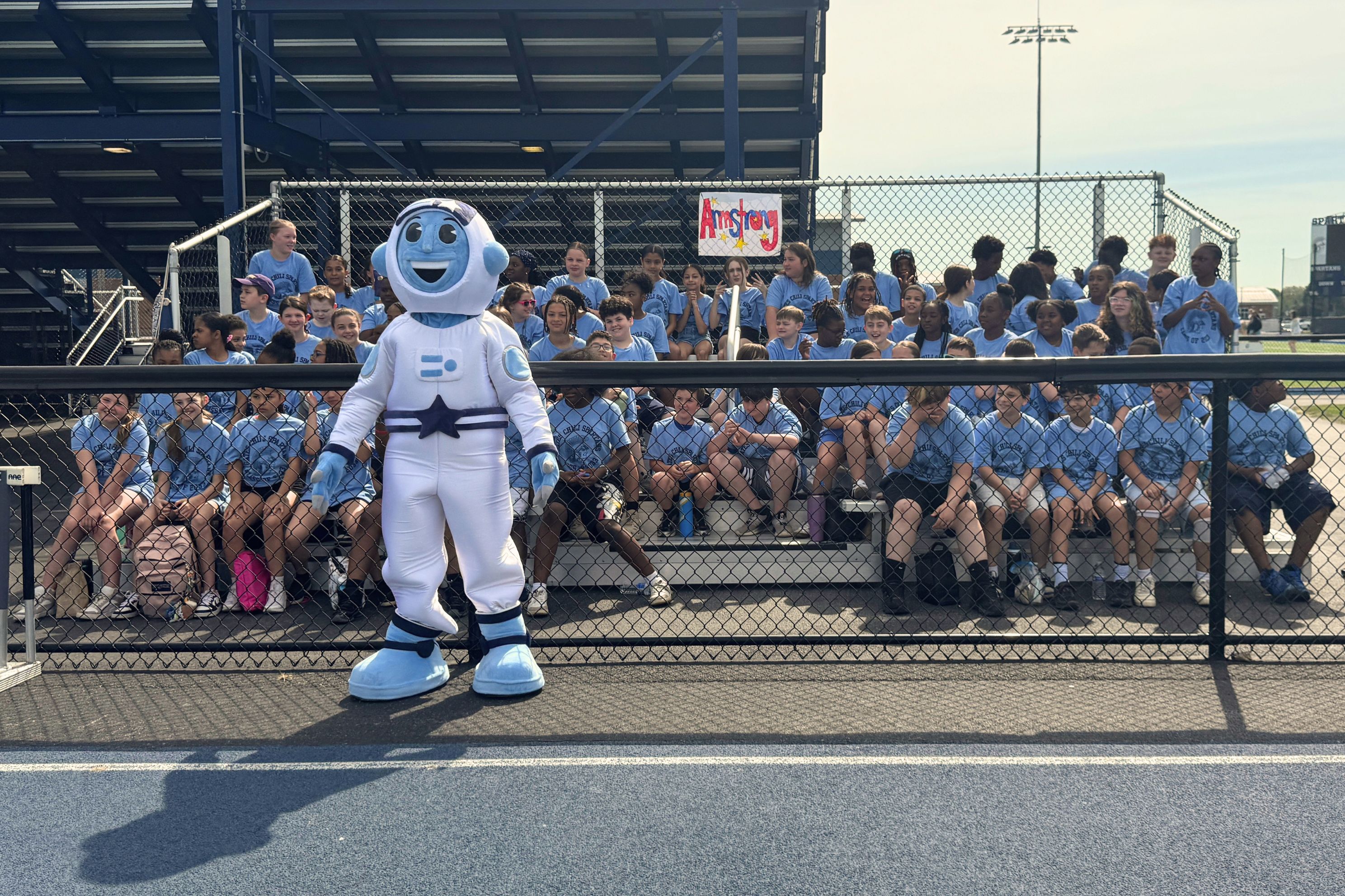A mascot in a white astronaut costume stands in front of a crowd of children in light blue shirts.