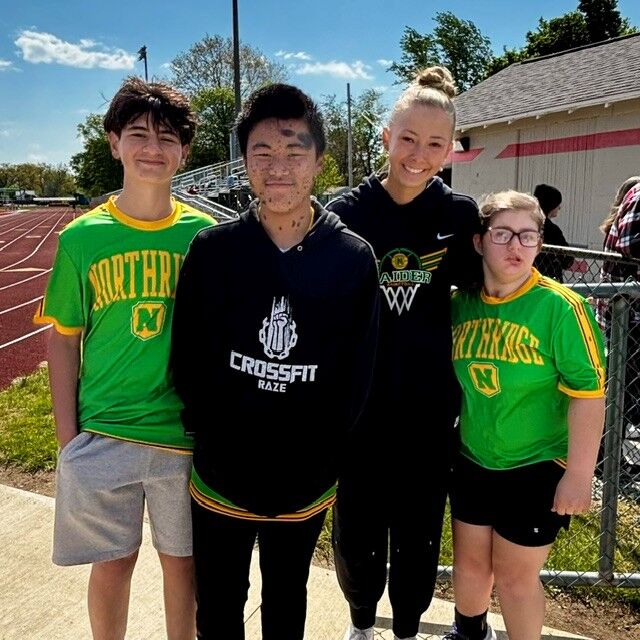 Four young people stand together outdoors on a sunny day, with a running track and bleachers in the background.