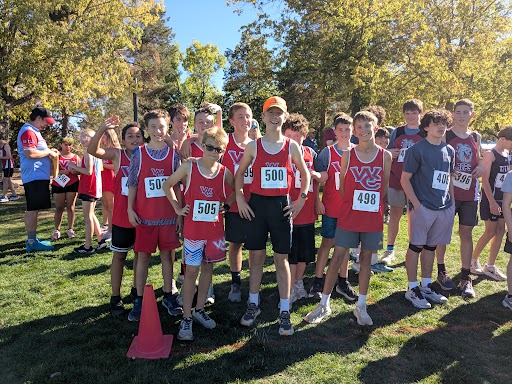 A group of young runners wearing numbered bibs gather together on a grassy field in the sunlight.