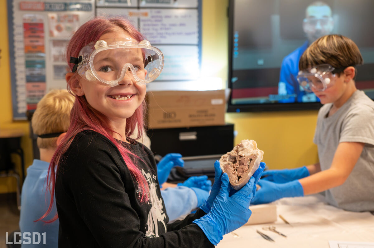 A smiling student wearing safety goggles and blue gloves holds an anatomical heart specimen in a science classroom.