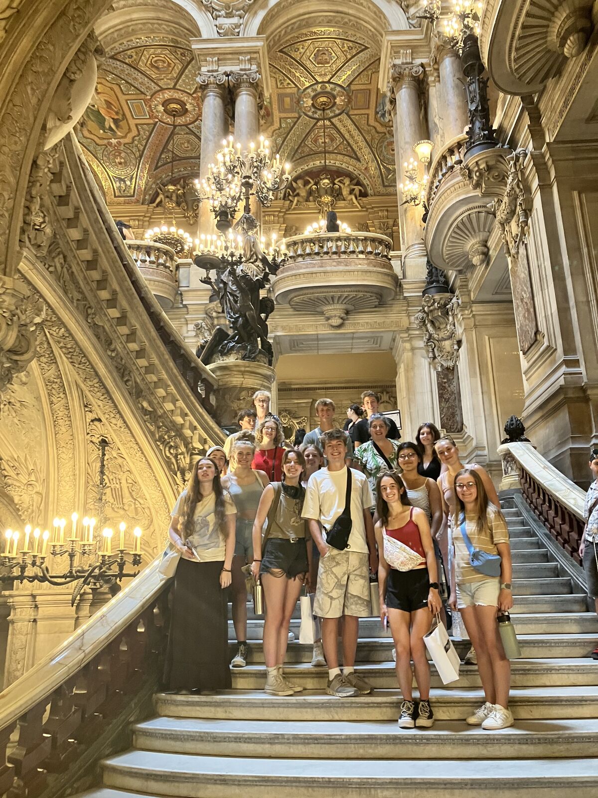 A group of people pose for a photo on a grand marble staircase inside an ornate, historic building with chandeliers and detailed ceilings.