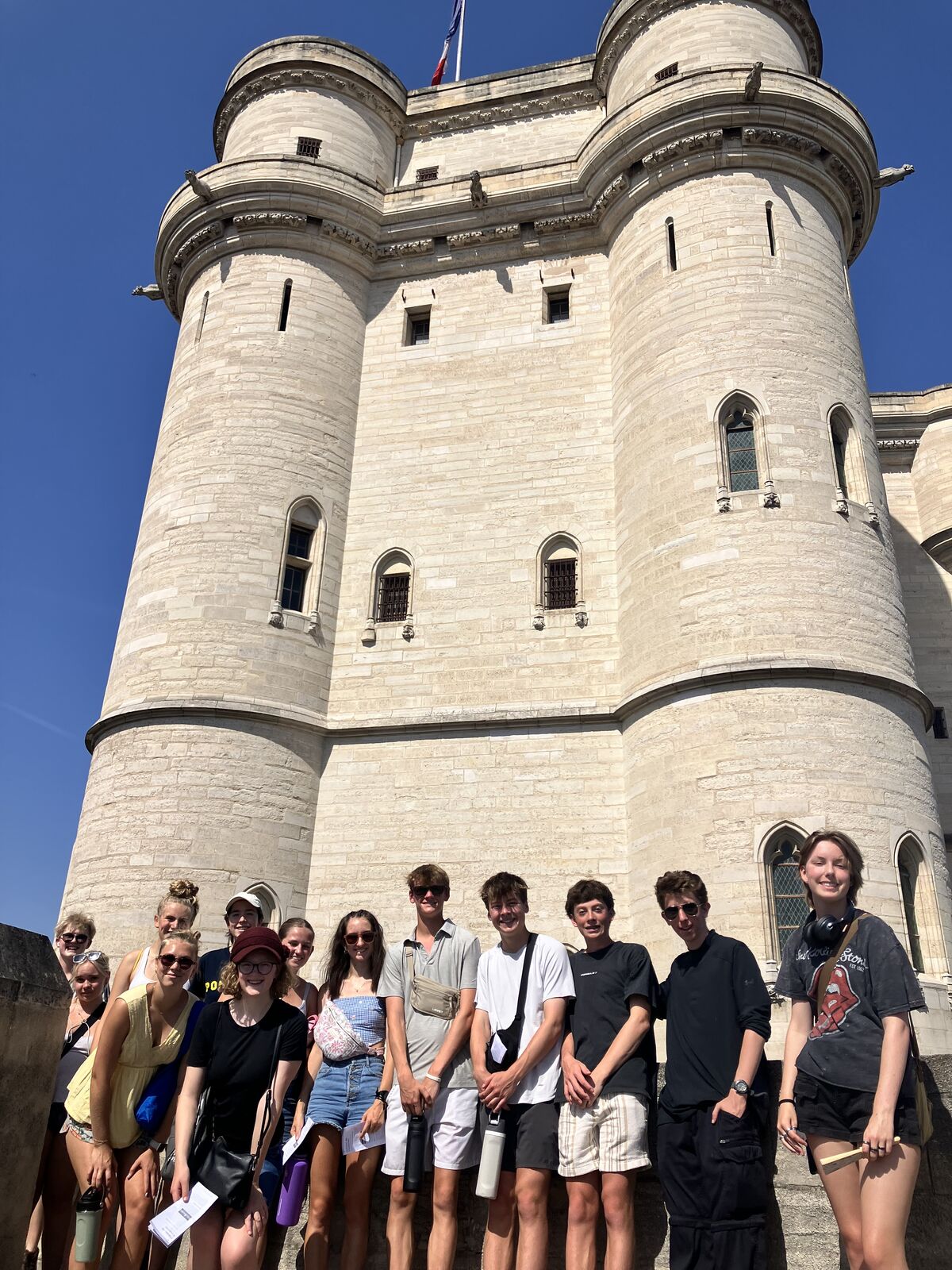 A group of friends stands in a line outside a large, historic stone castle with tall towers.