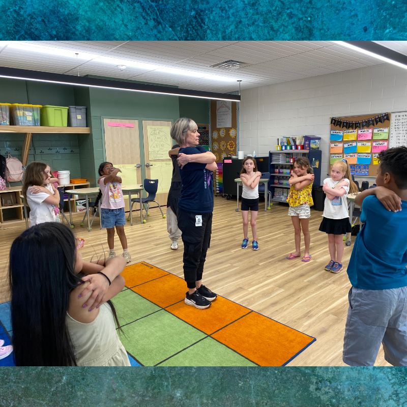 A teacher leads a group of children in a classroom exercise, with everyone crossing their arms.