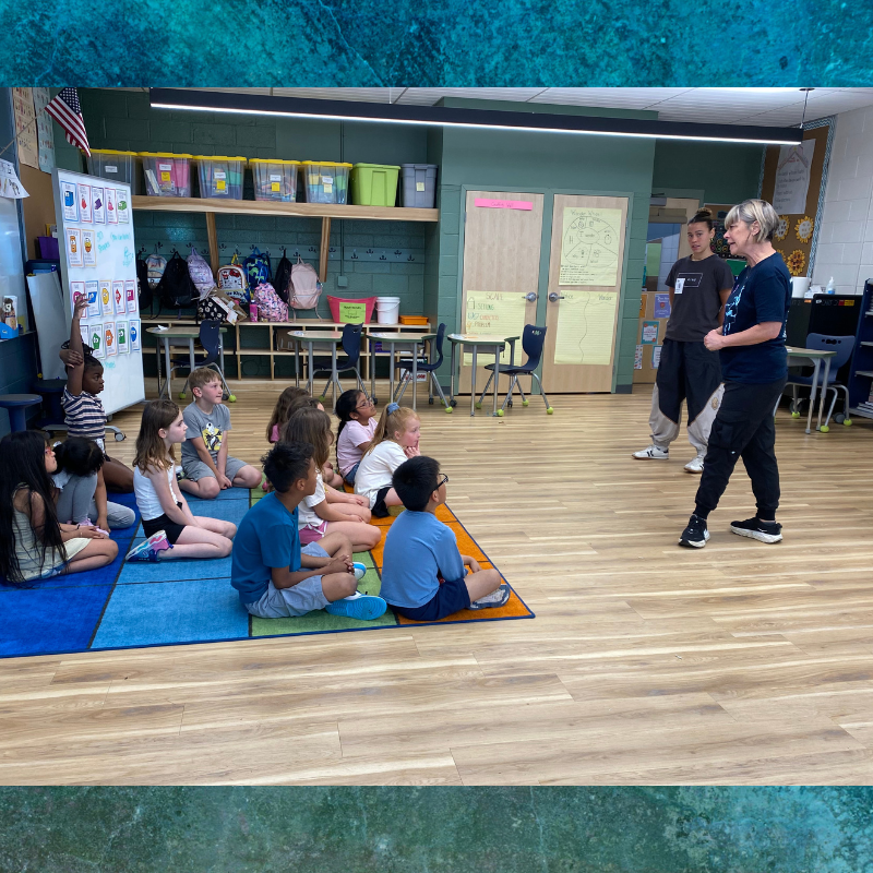 Children sit on a colorful rug in a classroom, listening to two adults.