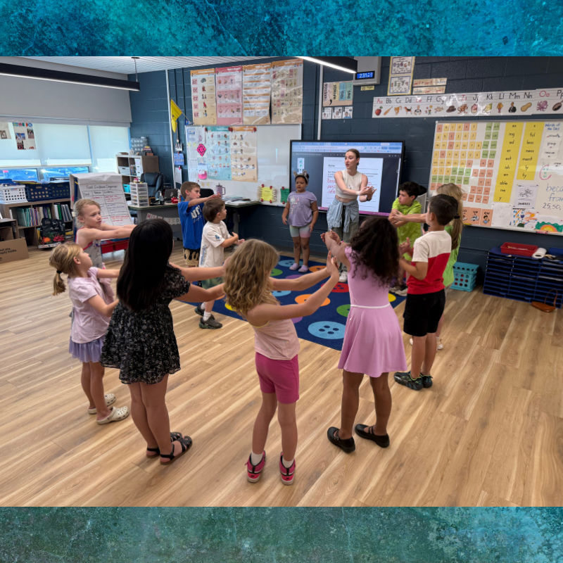 Children and a teacher hold hands in a circle in a classroom.