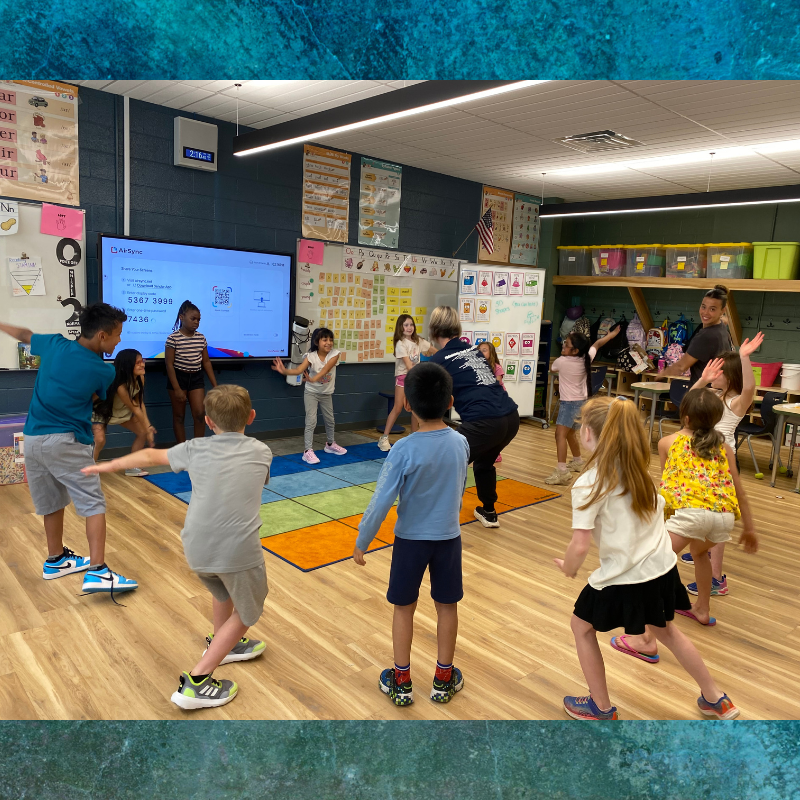 Children and an adult participate in an energetic activity in a brightly lit classroom.