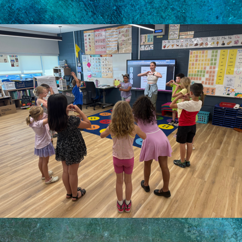 A teacher leads a group of young students in a classroom activity, with children mimicking hand gestures.