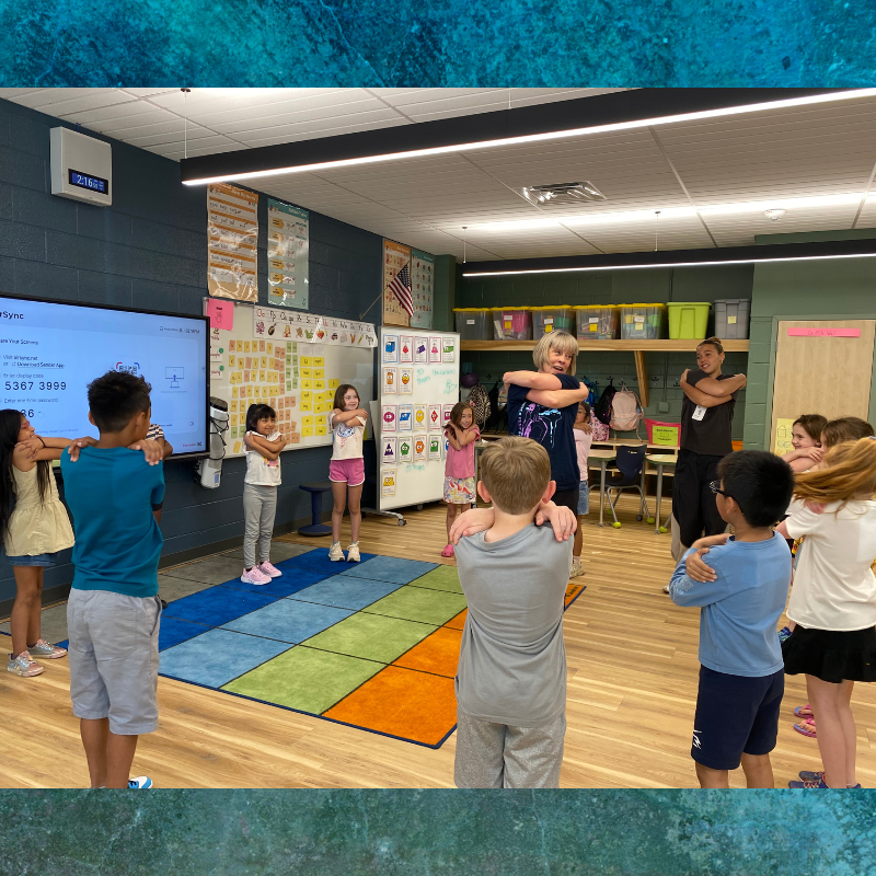 Children and adults participate in a stretching activity in a classroom with a large screen and whiteboard.