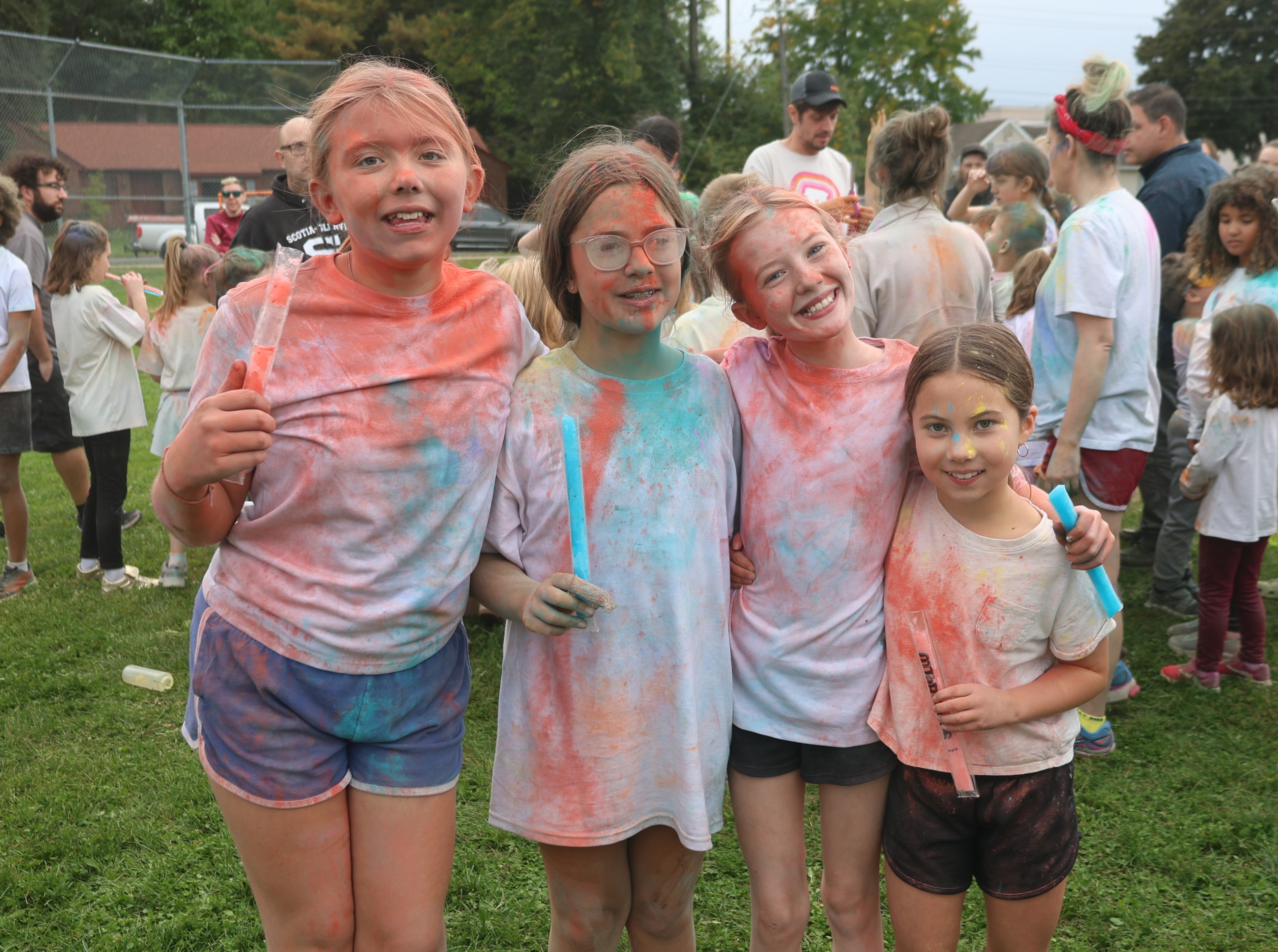 Four smiling young friends with colorful powder on their faces and clothes pose together outside.