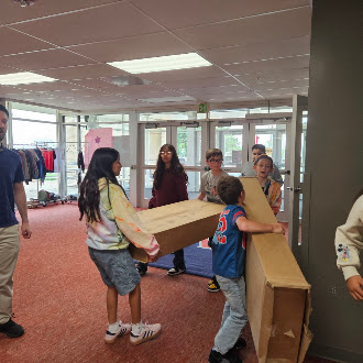 A group of students work together to carry long cardboard boxes inside a school building.