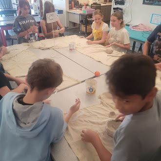 Students gather around a large table in an art classroom, working with clay.
