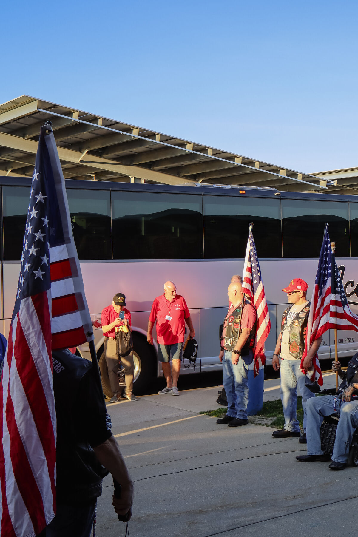 A group of people gather outdoors next to a large tour bus, holding American flags.