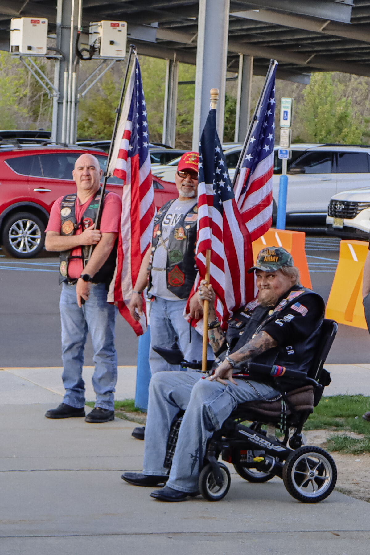 Three people holding American flags stand in a parking lot, with one person seated in a wheelchair.