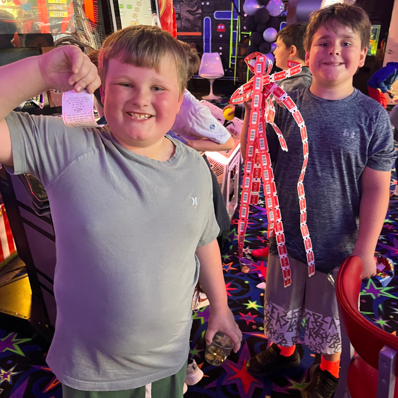 Two smiling boys stand in an arcade, one holding a ticket and the other a figure made of tickets.