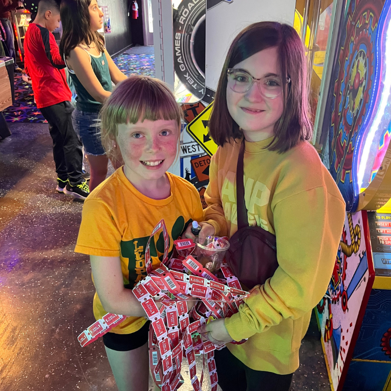 Two smiling girls hold a large bunch of red tickets in an arcade.