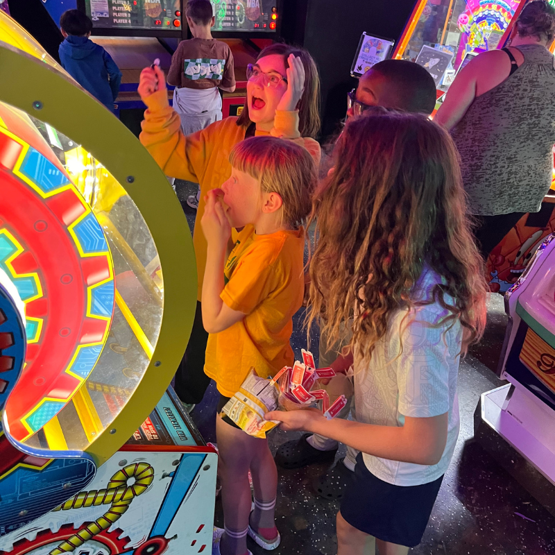 A group of children and adults are gathered around a brightly lit arcade game, with some looking excited and others focused on the game.
