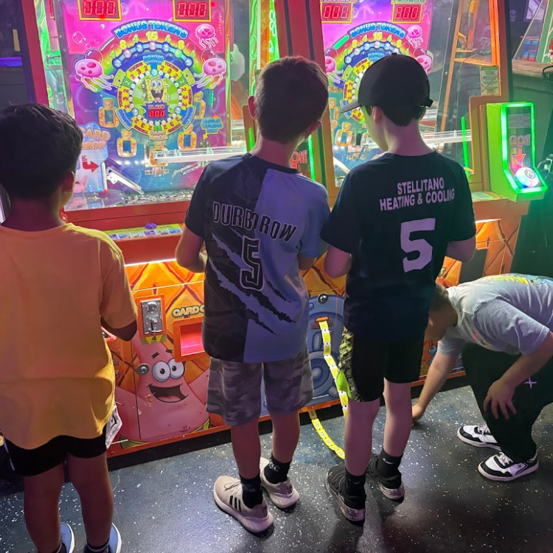 Three boys stand in front of brightly lit arcade games, one in a yellow shirt, another in a blue and black shirt, and the third in a black shirt.