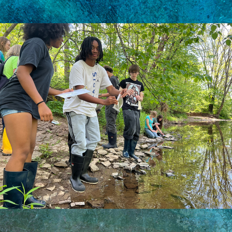 Students in boots examine a stream, holding a clear bag with water samples.